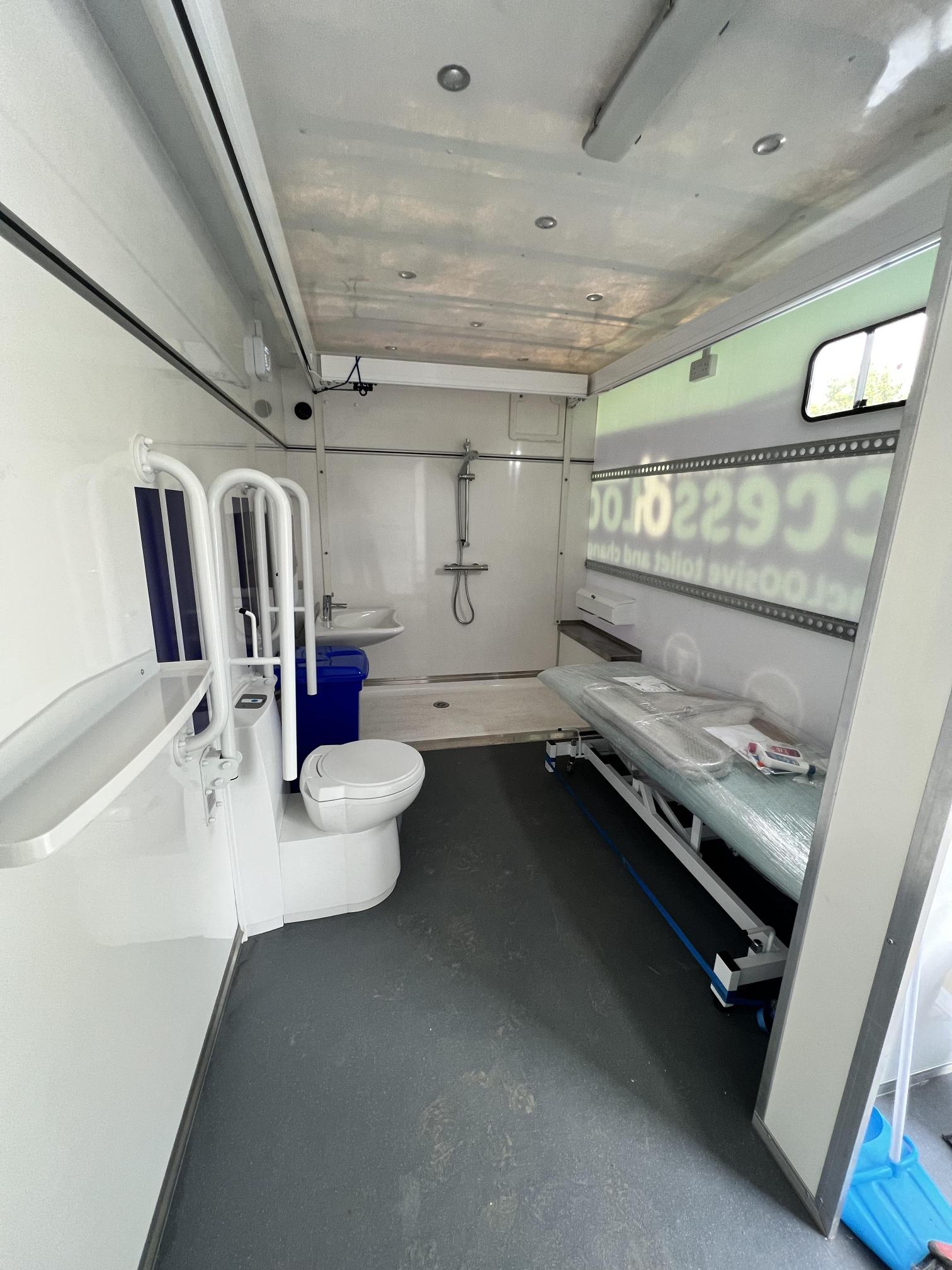 A view of the inside of the AccessoLoo with the changing table, lavatory, sink, shower and ceiling hoist in view.