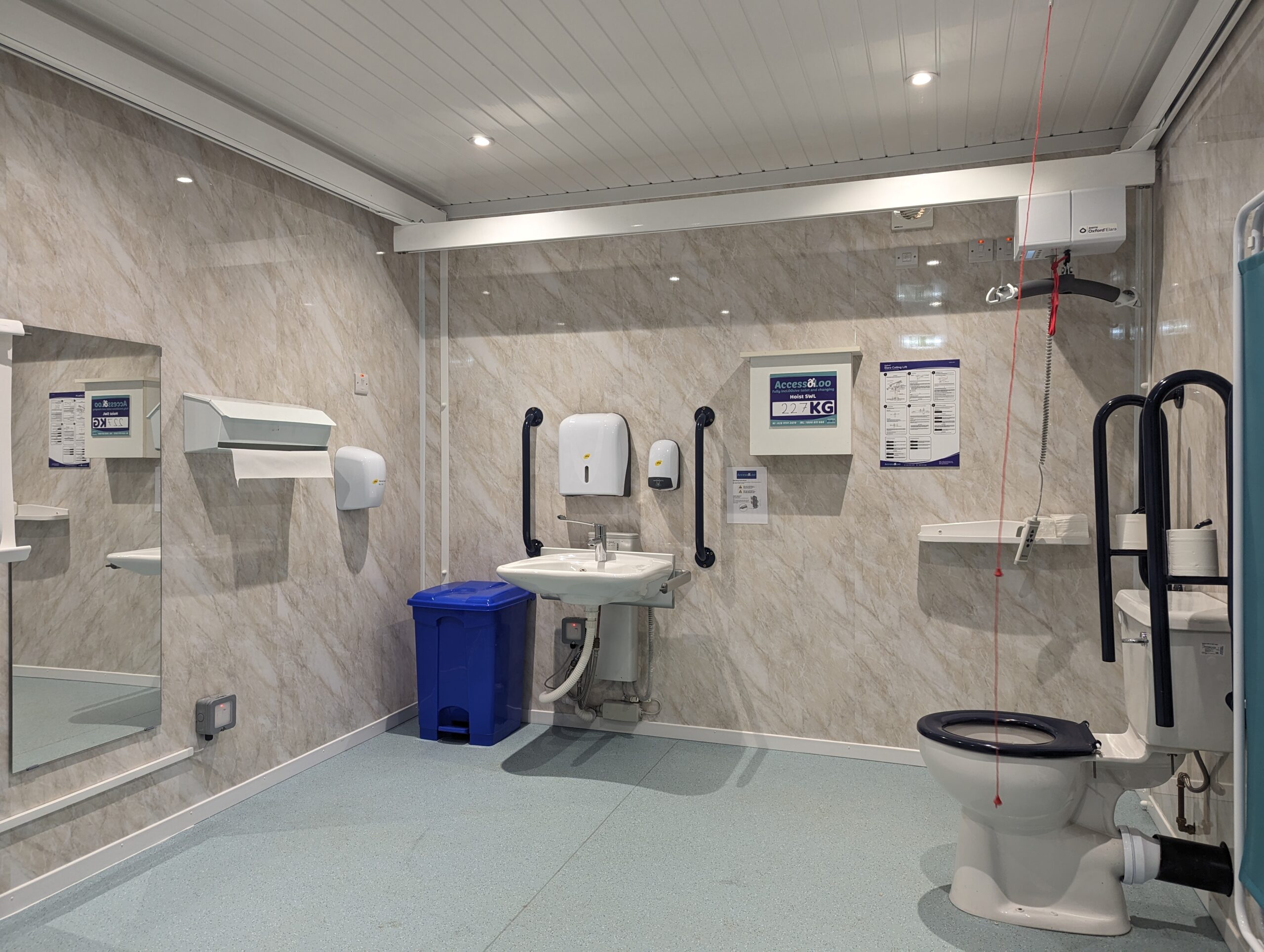A view of inside a modular Changing Places with toilet, sink, ceiling hoist, mirror and litter bin in view.